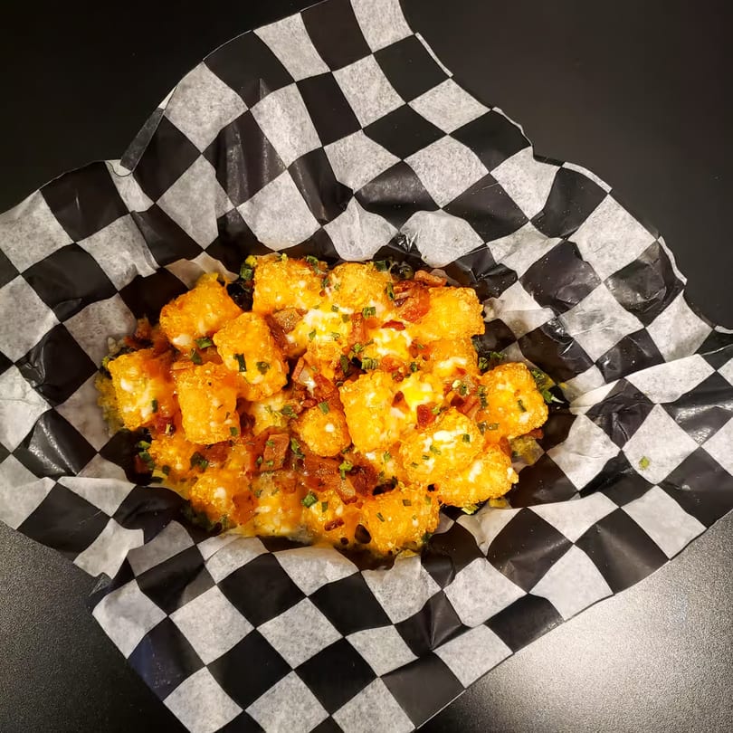 Loaded tater tots with melted cheese, bacon bits, and chives in checkered paper.