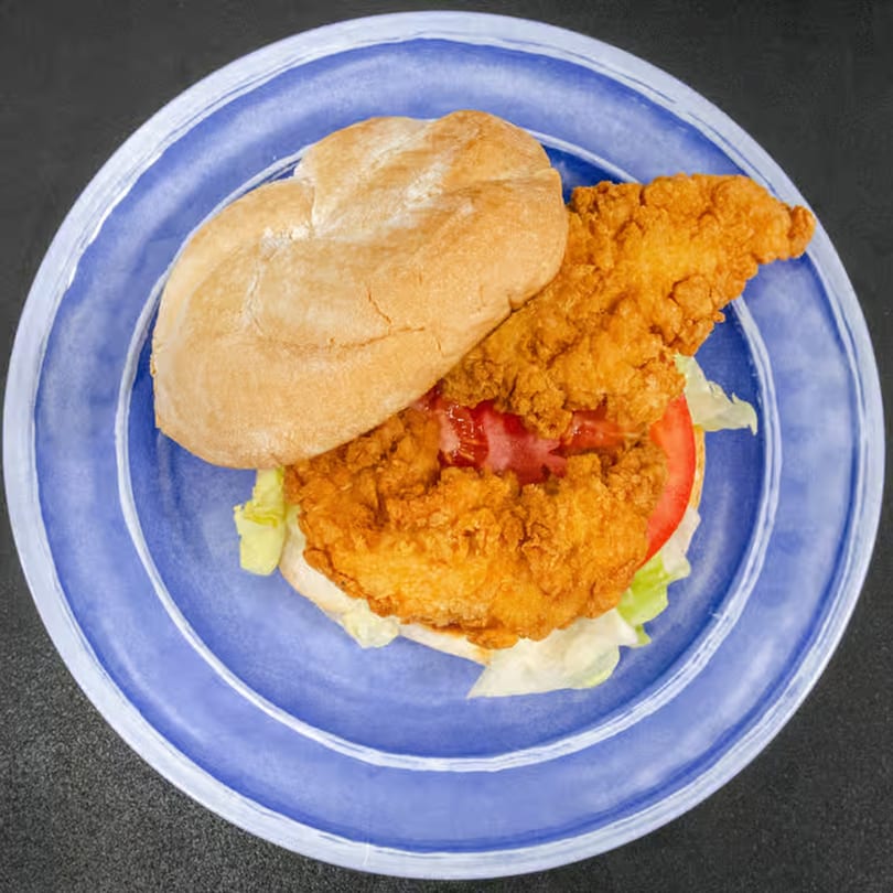 Crispy fried chicken sandwich with lettuce, tomato, and ketchup on a bun, served on a blue plate.